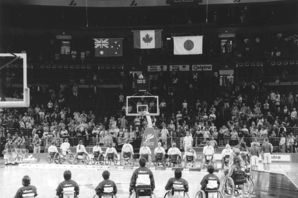 22 wheelchair users line of on a basketball court to receive a Paralympic medal. A crowd of spectators watch on from the stands. 