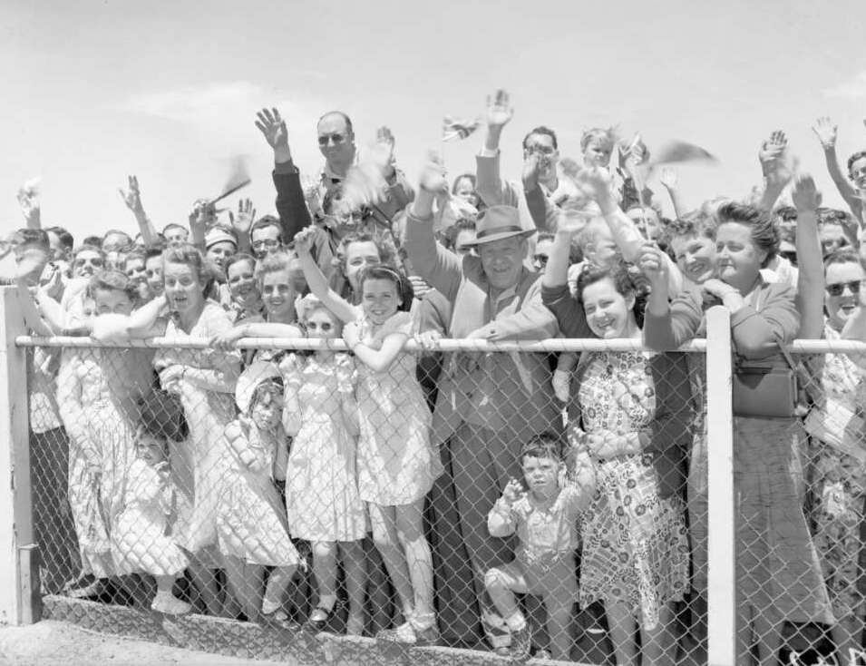 A large crowd of men, women and children stand behind a fence. They are all happy and smiling and many are waving.