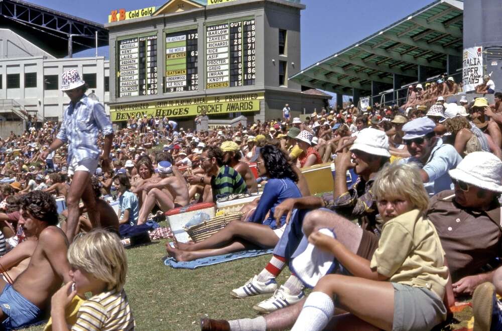 A crowd of spectators sit on a grassy slope watching a cricket match happening off camera
