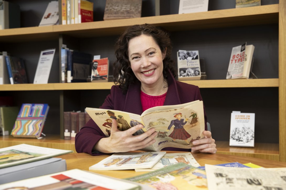 Woman with black curly hair smiling as she holds open an old book about wool and knitting. More similar books are spread out on the table in front of her, and books on various topics sit on the shelf behind her.