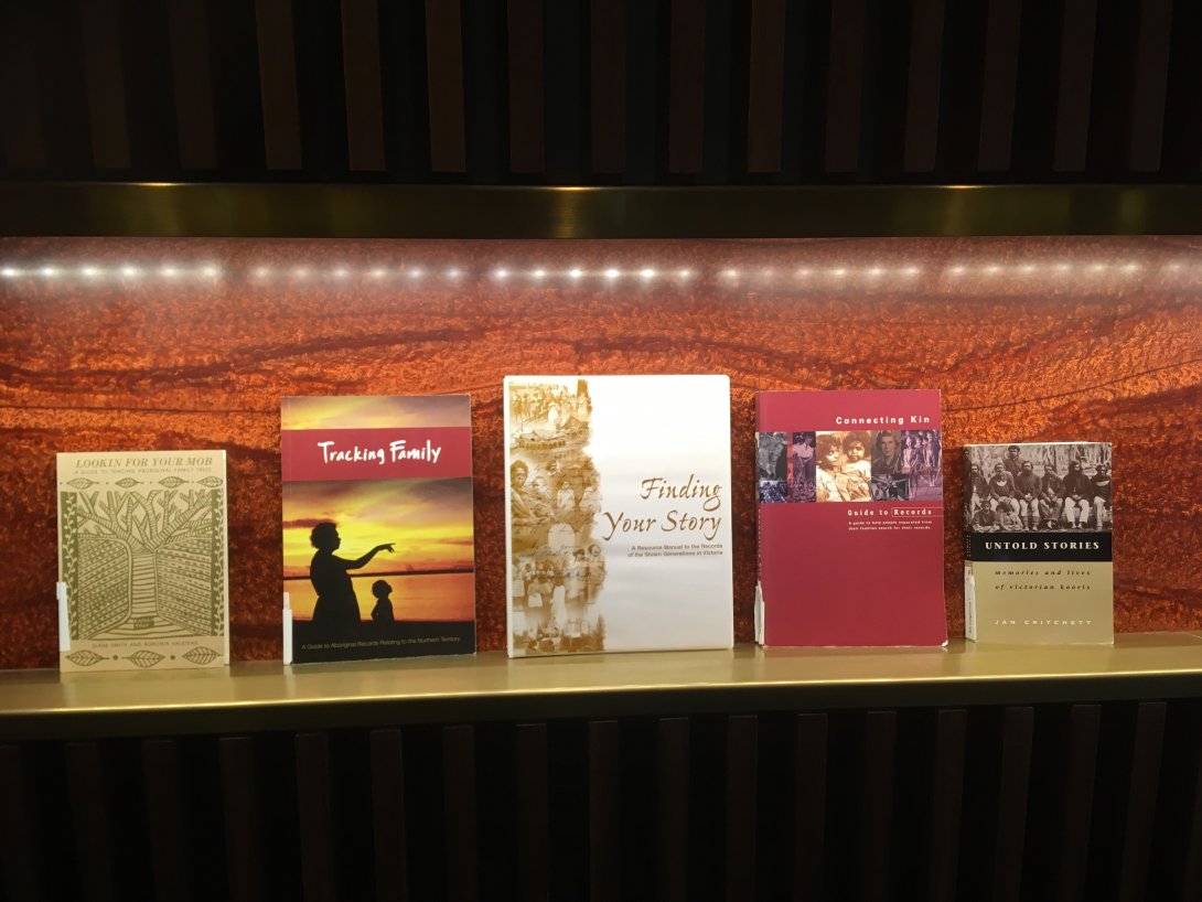 Five reference books about First Nations family history of various sizes on a lit shelf, including one book titled 'Tracking Family' which has a pho of a woman and child looking at a sunset and another titled 'Finding your story'