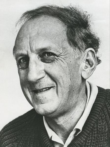 Black and white portrait photo of old man smiling. He wears a knit jumper over a collared shirt