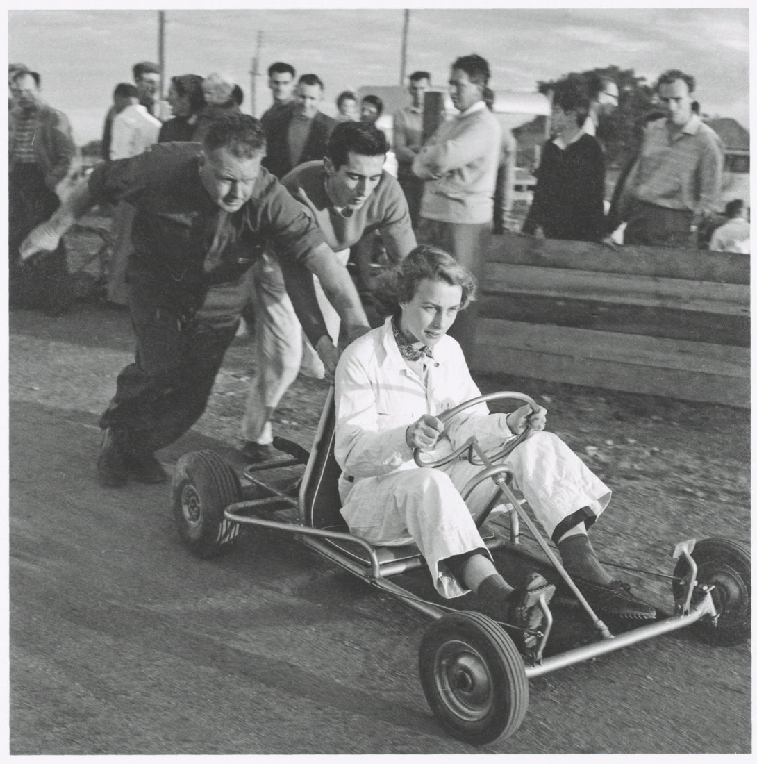 Two men pushing a go-kart with a woman in the drivers seat. Black and white image.