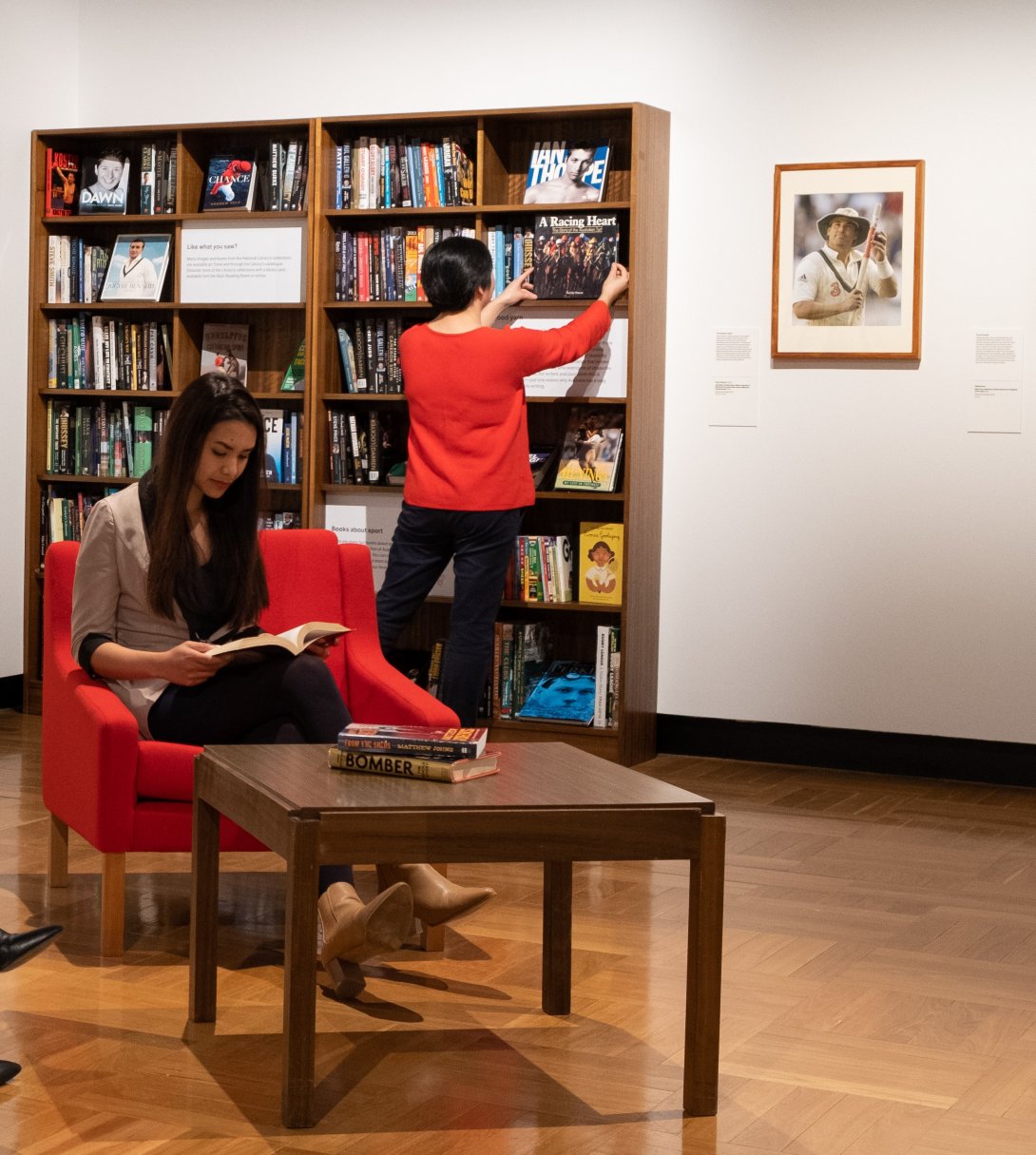 Woman sitting on a red armchair reading in front of large bookshelf with another woman looking at a book placed on the shelf beside a framed image of Shane Warne