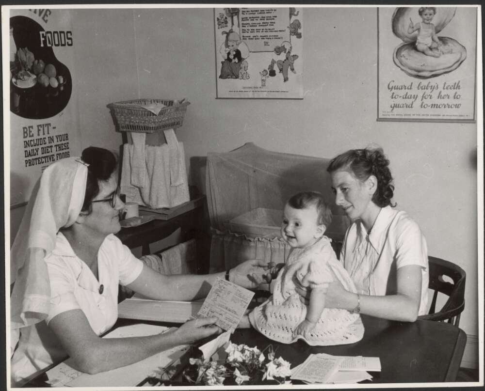 Nurse attending to baby and mother. Mother and nurse sit in chairs by a table while baby is being held so it sits on the table.  All three are smiling.