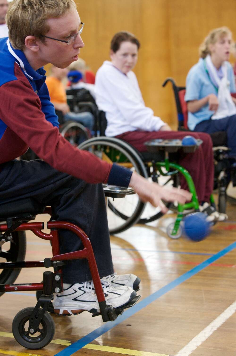 Man in wheelchair participating in game of boccia, with his hand outstretched and the ball in midair. Two women in wheelchairs sit behind him