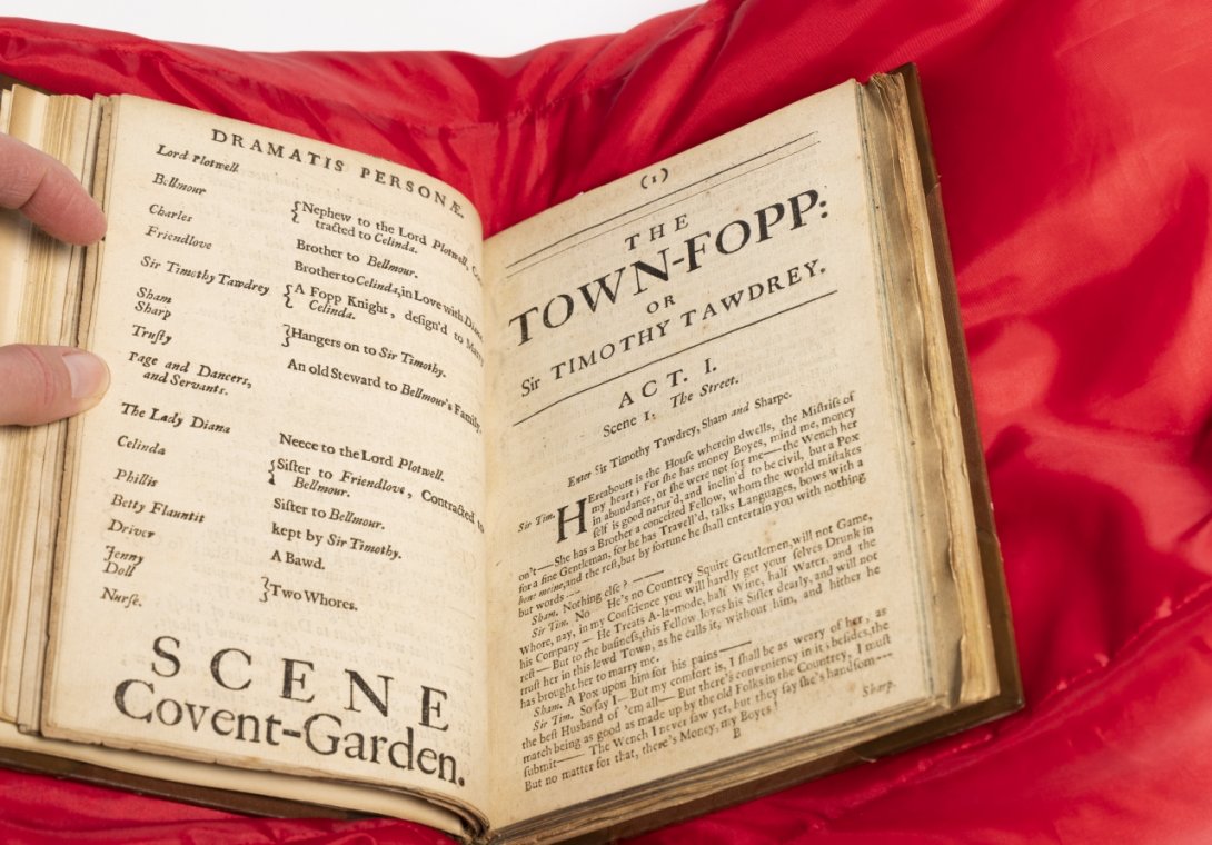 Photo of a rare book on a red cushion open to the beginning of a play with cast list on the left