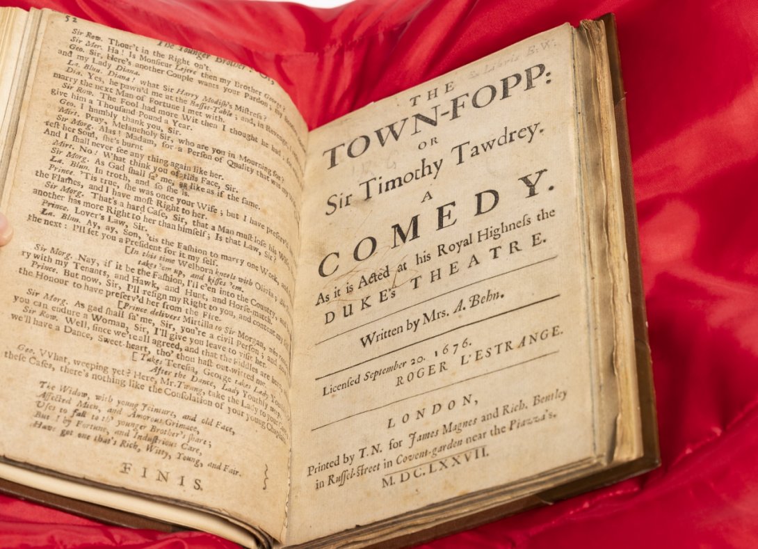 Photo of a rare book on a red cushion with large font text (of a play) on the right hand side