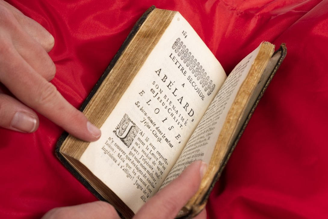Photo of a rare book on a red cushion being held open by two hands.