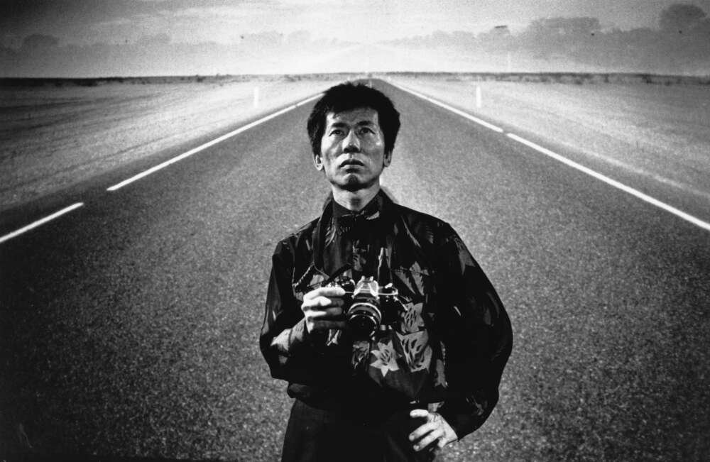Chinese-Australian man in black shirt standing on an empty road holding a camera with a hand on his hi[