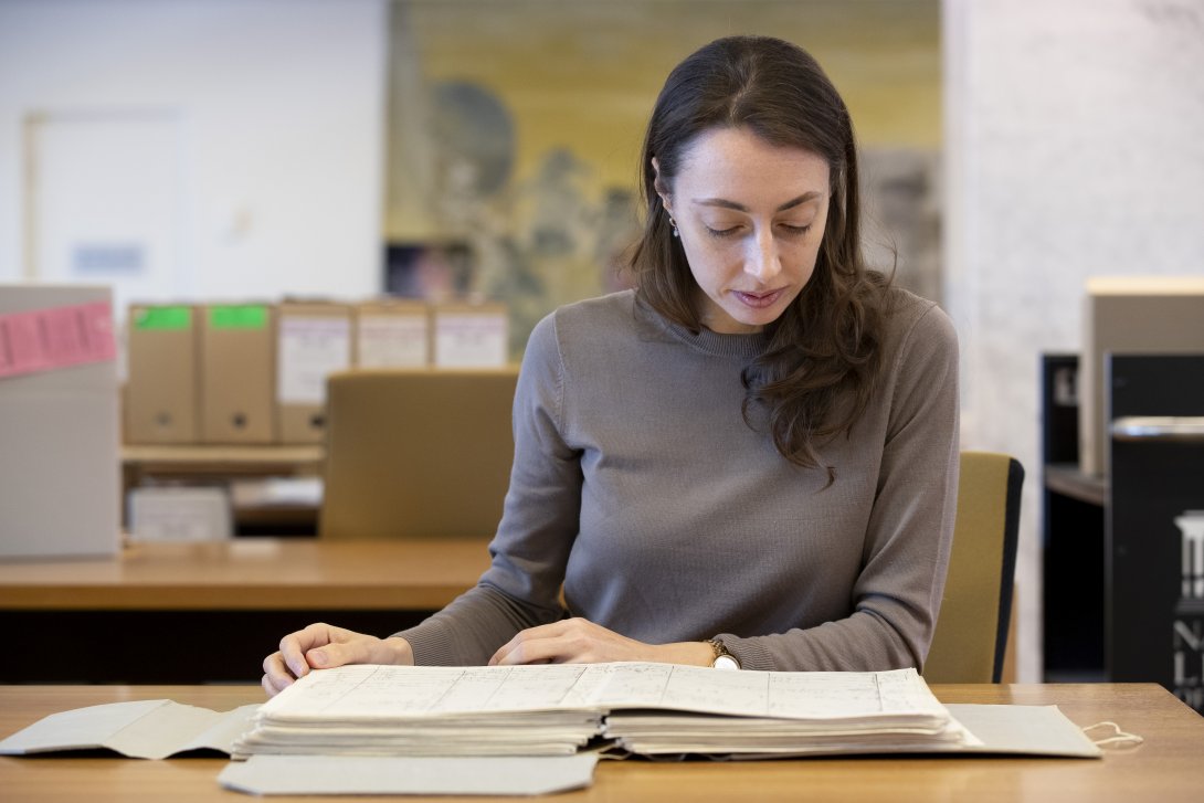 Woman with brown hair and eyes, wearing a brown sweater, sitting at a desk in the Special Collection Reading Room, looking down as she reads from an old book.