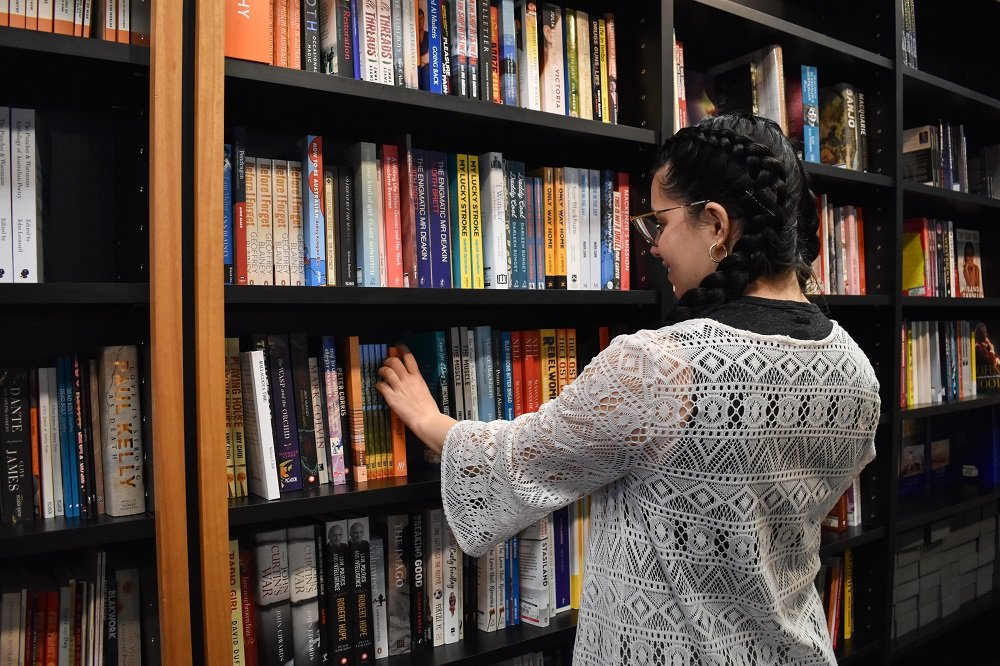 Young women with dark hair in braids and a lace long-sleeved shirt browsing the shelves of a bookshop and reaching out to pick up a book