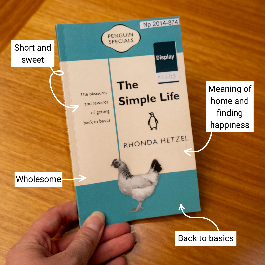 Book with blue and white cover with a chicken on it and text reading 'The Simple Life' and 'Rhonda Hetzel' being held up in front of a wooden background. Annotations around the book read 'short and sweet', 'wholesome', 'meaning of home and finding happiness' and 'back to basics'.