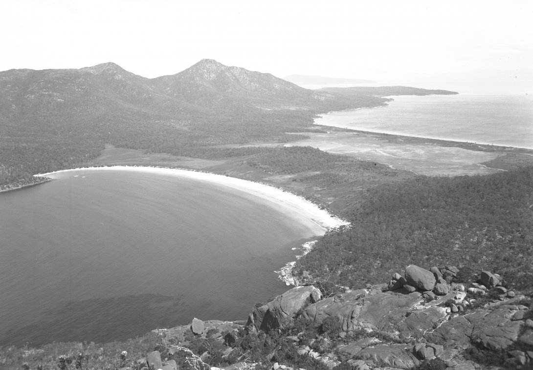 Black and white photograph of two beaches/bays on either side of a strip of land. The land between the bays in relatively flat, while on each end are mountains and forest