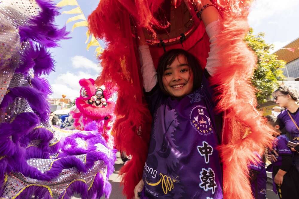 Young child in purple shirt smiles as she holds her lion costume for the Moon Festival above her head