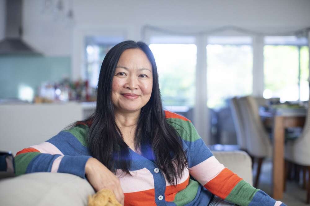 Woman of Chinese heritage sitting on a couch in a nice house, wearing a colourful striped jumper and smiling