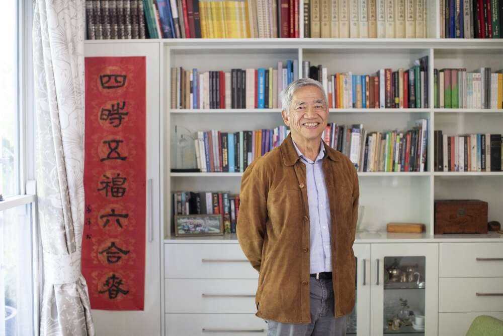 Older man of Chinese heritage in a brown jacket smiling and standing in front of a neat, colourful bookshelf