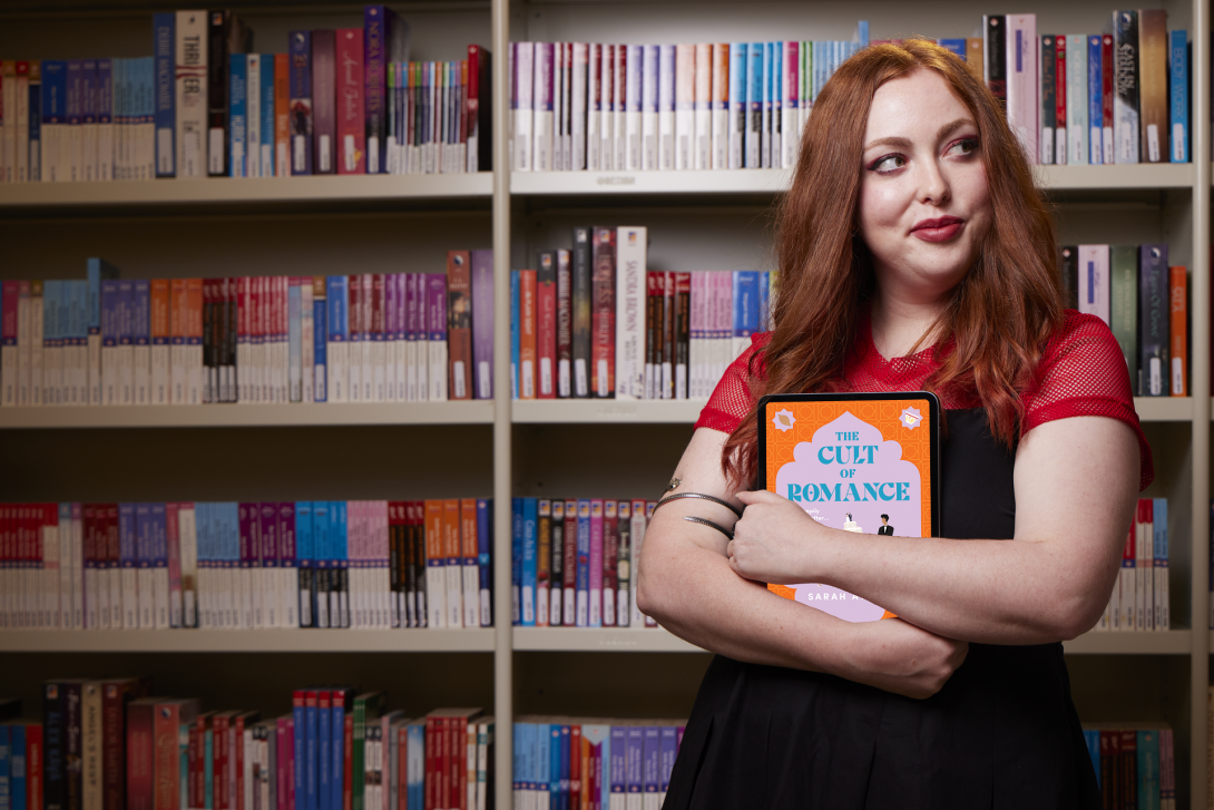 Woman with red hair standing in front of a bookshelf of romance books, holding a tablet with the cover of a romance book on the screen. She is facing forward with her back to the bookshelf, looking to the side and slightly smiling