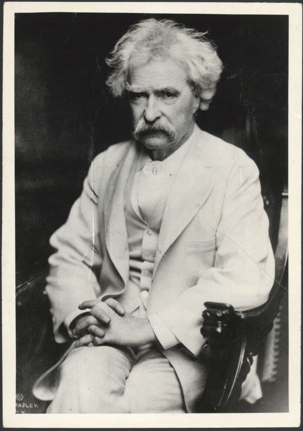 A black and white photograph of a man wearing a white suit. He is sitting down and also has a moustache.