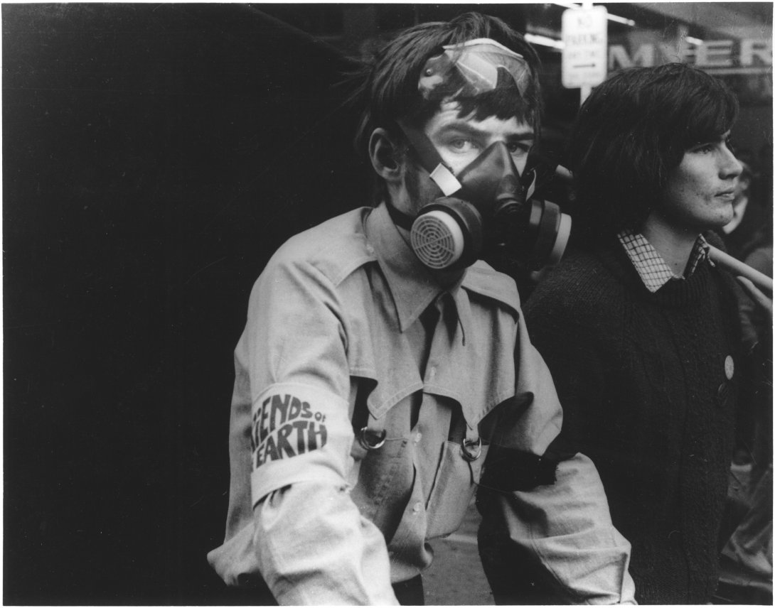 Young man, maybe late teens, wearing a grey shirt with a 'Friends of the Earth' armband wearing a gas mask