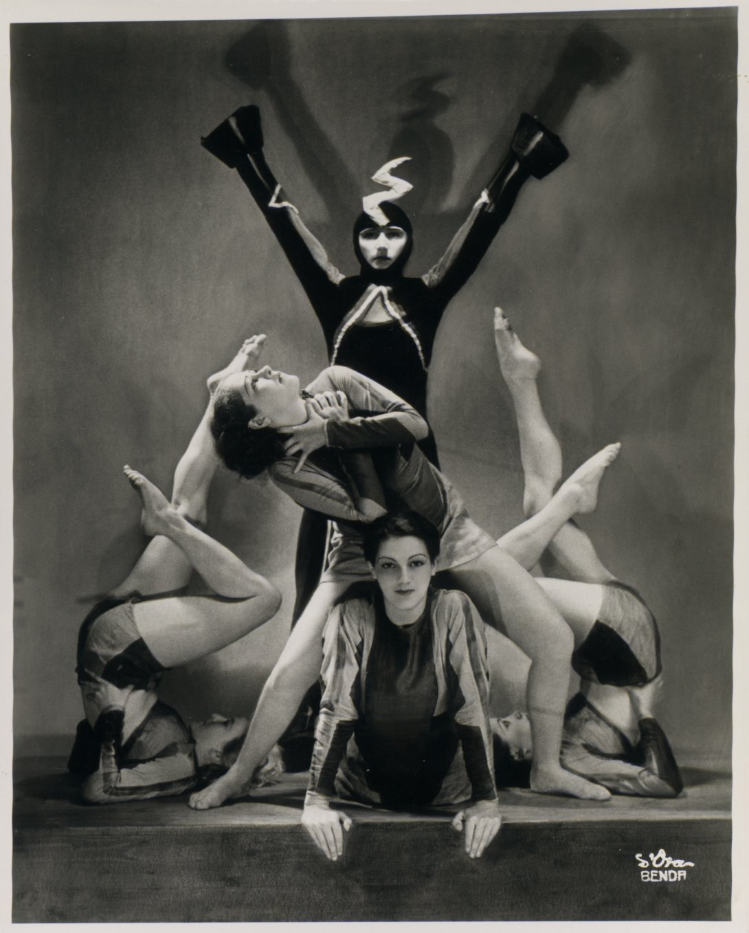 Five dancers in various costumes posing. Two dancers are balancing on their backs with their legs in the air, one is standing over the shoulders of another, and one at the back has their arms up.