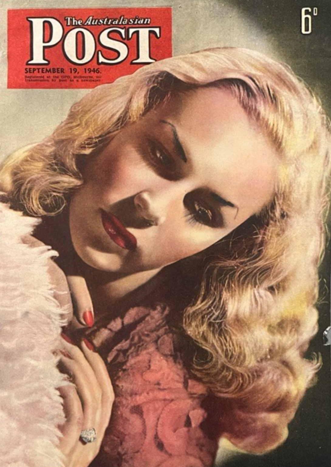 Cover of a magazine with a blonde woman in view form the shoulders up. She appears to be leaning around a pink fluffy cushion, and is wearing a red/pink shirt, red nail polish and a diamond ring