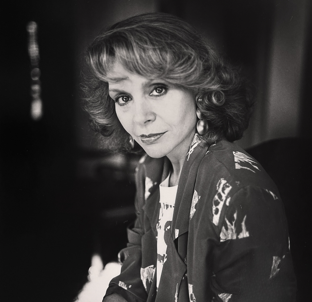 Black anf white photograph of a woman with light, short curly hair looking directly into the camera with a small smile. She's wearing a dark blazer with light, abstract shapes on it and large round earrings