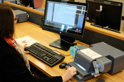 A woman sits at a desk viewing microfilm at a computer monitor